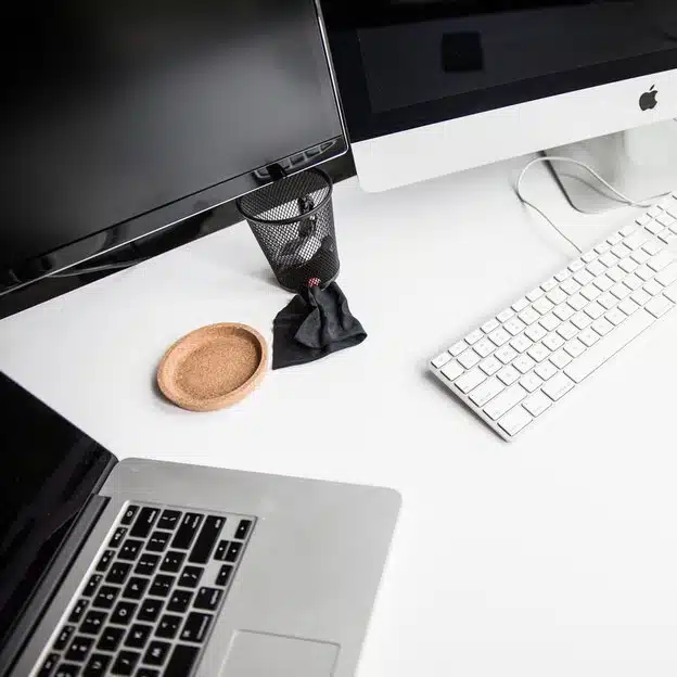 A tidy workspace with a Mac laptop, two monitors, and a keyboard on a white desk. A cork coaster and a metal mesh pen holder add minimalist accents.